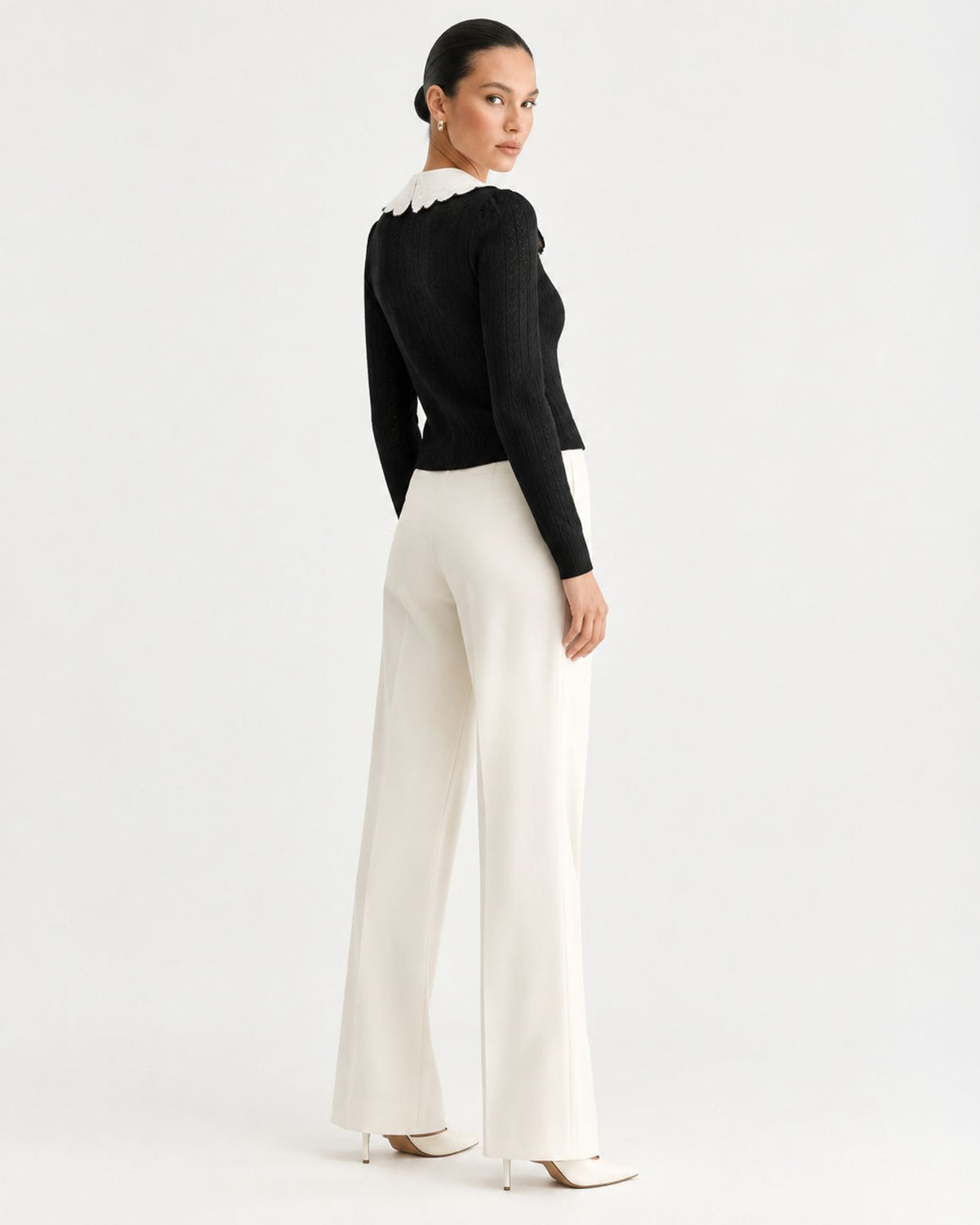 A model is shown from the back wearing a black long-sleeve knit top with a white ruffled collar and high-waisted, wide-leg off-white trousers, paired with white pointed-toe heels, against a plain white studio background.