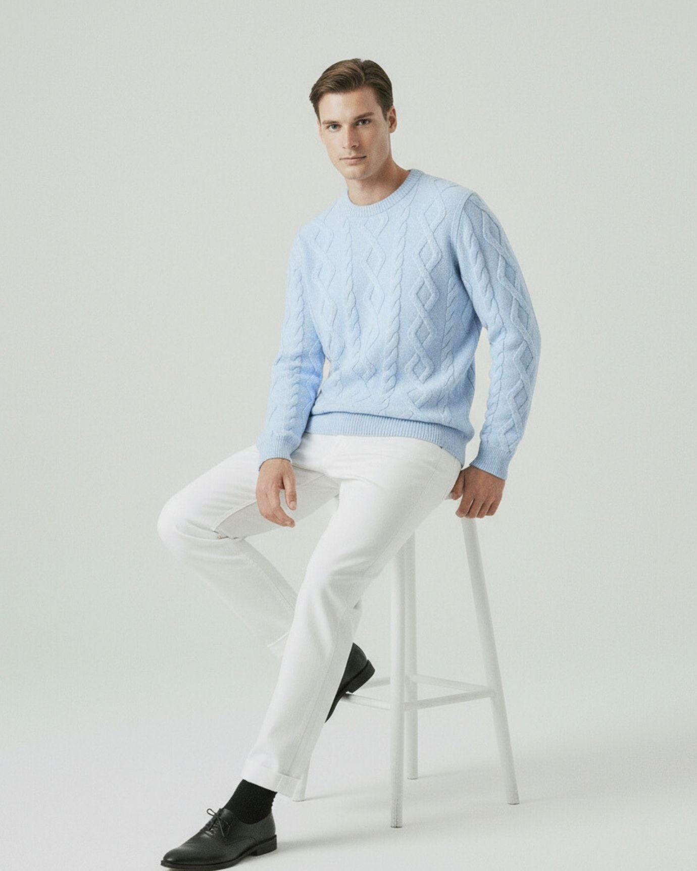 Man wearing a light blue cable knit sweater and white pants sitting on a stool against a plain background