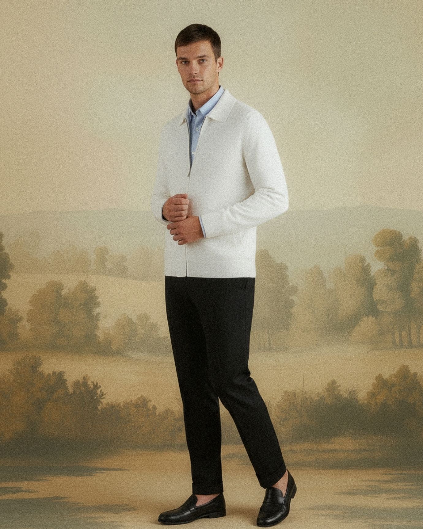 Man wearing a white sweater over a blue shirt with black pants and shoes, standing against a scenic wall.